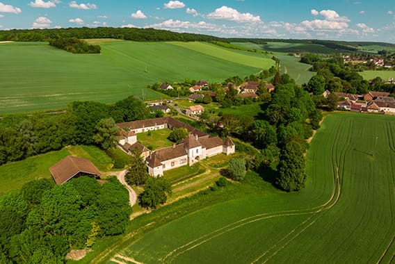Château des Roises à Bucey-en-Othe 7 - © Studio OG Vue aérienne d’un village rural entouré de champs verdoyants et de collines en campagne française, avec des maisons en pierre et une ferme au cœur d’un paysage agricole sous ciel bleu